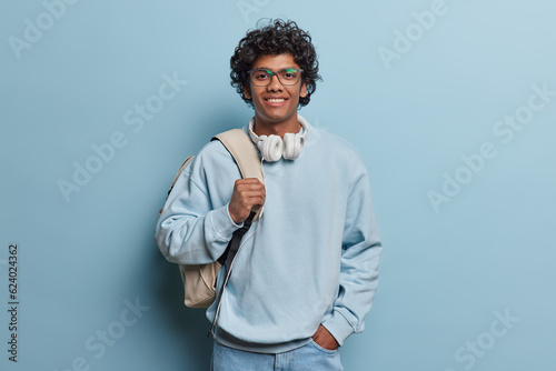People lifestyle concept. Studio shot of young happy smiling Hindu male student standing isolated in centre on blue background wearing casual hoodie and jeans with white leather bag on left shoulder