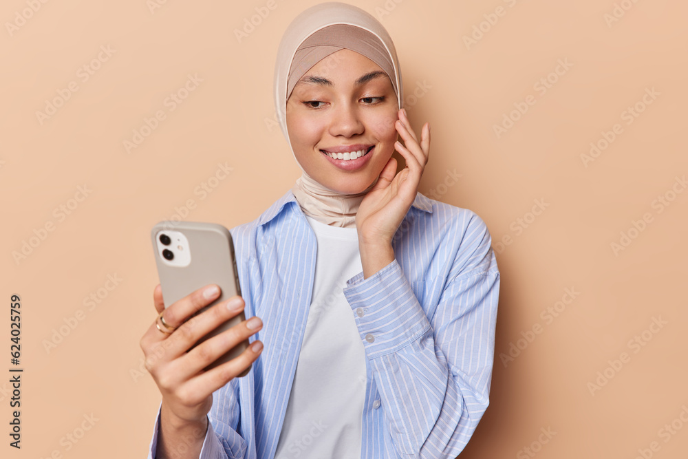 Happy tender Islamic woman touches face gently focused at smartphone smiles pleasantly dressed in formal clothing and veil on head isolated over brown background. People religion technology concept
