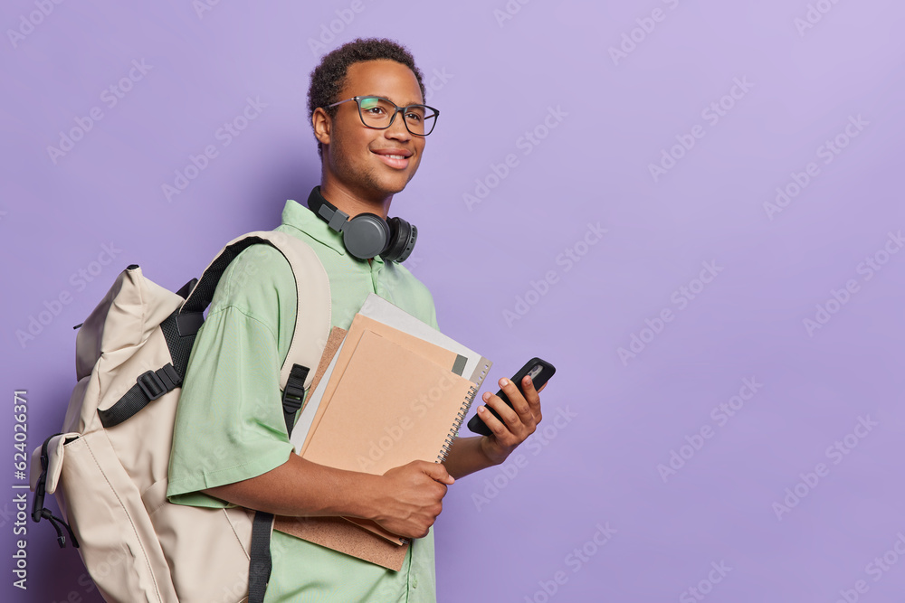 Studio shot of young happy smiling African student wearing green casual tshirt with big white bag holding books notebooks and smartphone standing on left with blank space for your advertisement Studio shot of young happy smiling African student wearing green casual tshirt with big white bag holding books notebooks and smartphone standing on left with blank space for your advertisement