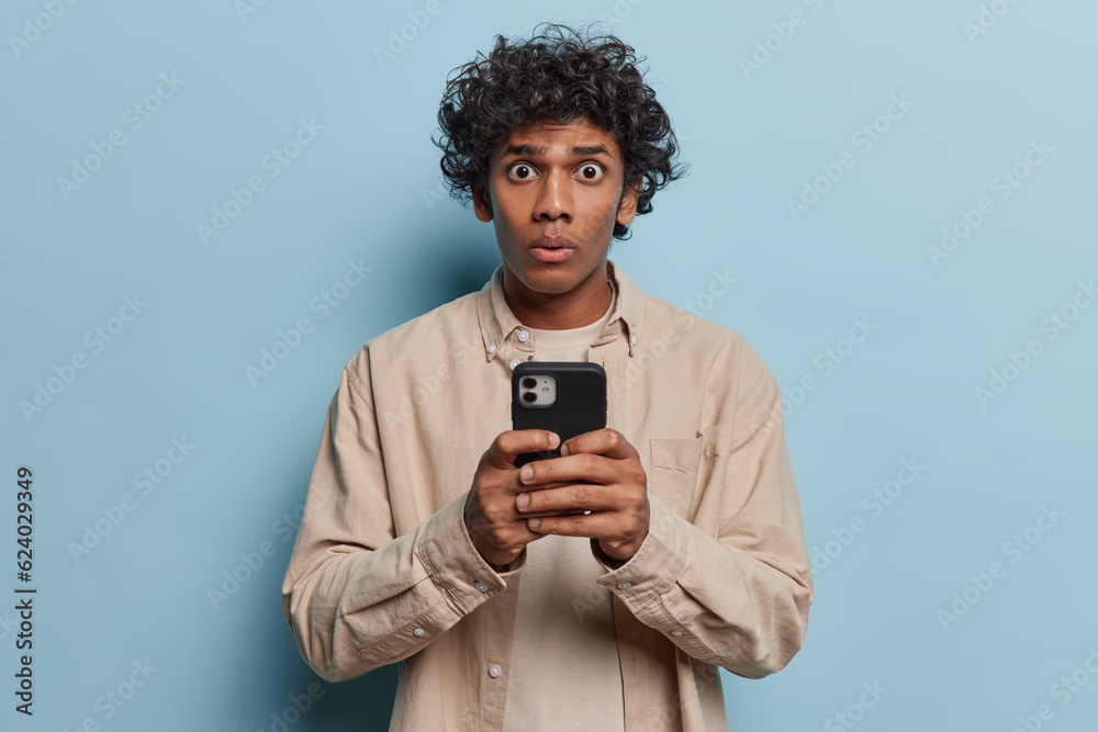 Horizontal shot of shocked Hindu teenage guy holds mobile phone reads shocking message from friend forgot about important meeting wears beige shirt isolated over blue background. Big online sales Horizontal shot of shocked Hindu teenage guy holds mobile phone reads shocking message from friend forgot about important meeting wears beige shirt isolated over blue background. Big online sales