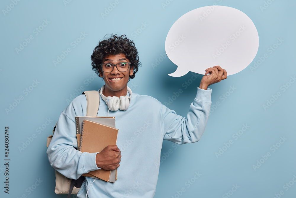 Smiling young happy Hindu male wearing casual hoodie with leather bag and headphones standing isolated on blue background holding books and white speech bubble for your promotion.Education concept Smiling young happy Hindu male wearing casual hoodie with leather bag and headphones standing isolated on blue background holding books and white speech bubble for your promotion.Education concept
