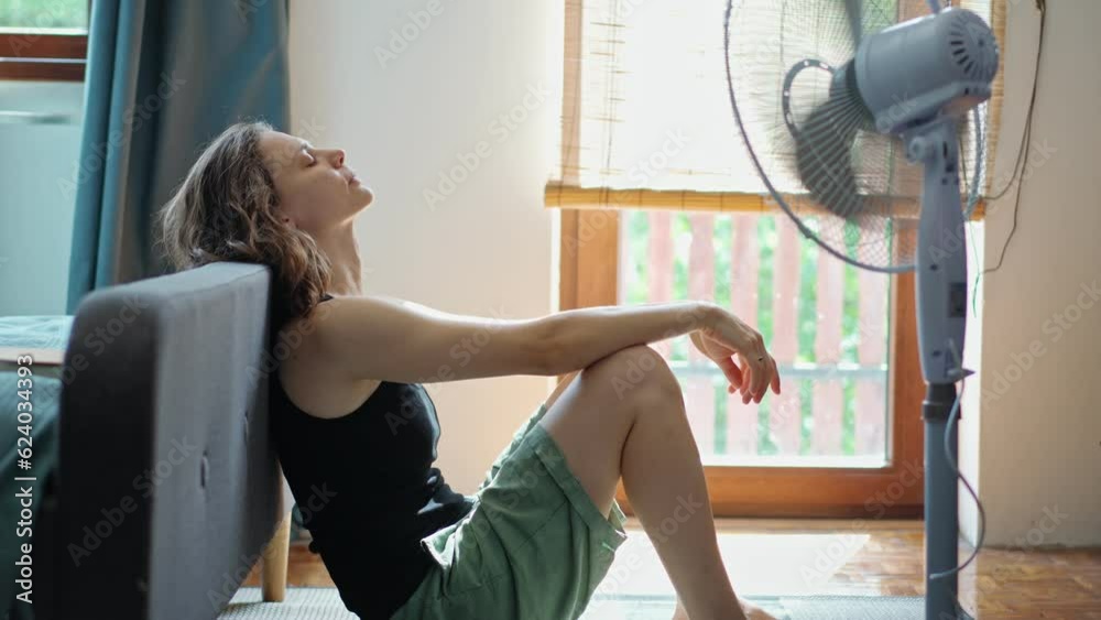 A young Caucasian woman tired from the summer heat sitting on the floor in front of a working electric fan.