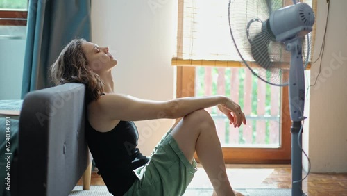 A young Caucasian woman tired from the summer heat sitting on the floor in front of a working electric fan.