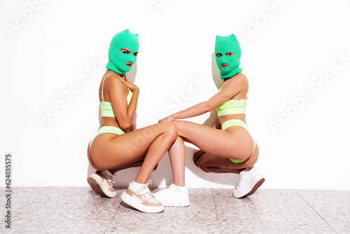 Two beautiful sexy women in green underwear. Models wearing bandit balaclava mask. Hot seductive female in nice lingerie posing near white wall in studio. Crime and violence. Sits on floor