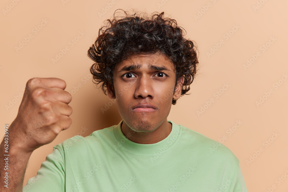 Angry discontent Hindu man with dark curly hair clenches fist expresses annoyance being outraged dressed in casual green t shirt isolated over brown background. I will show you how to offend me Angry discontent Hindu man with dark curly hair clenches fist expresses annoyance being outraged dressed in casual green t shirt isolated over brown background. I will show you how to offend me