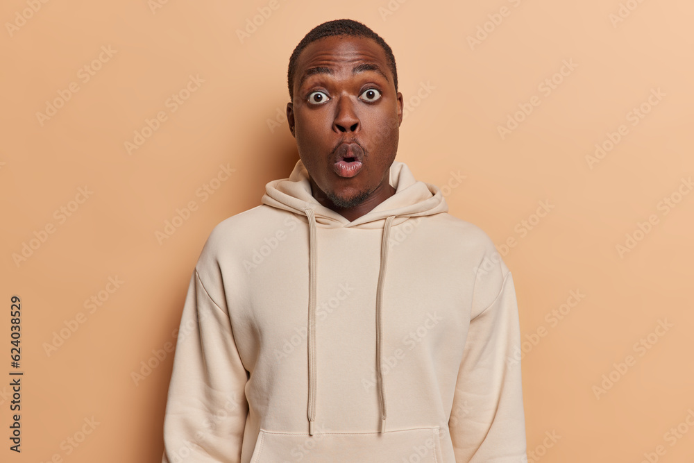 Attractive dark skinned man witnessing something truly extraordinary evoking sense of wonder and shared exhilaration stands speechless dressed in casual sweatshirt isolated over brown background