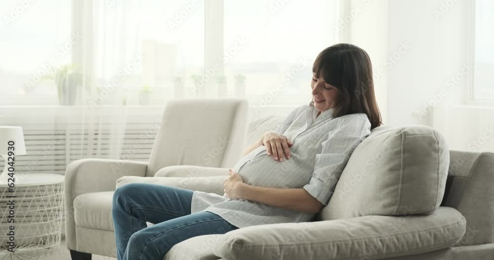 A pregnant woman sits on the living room couch, her face beaming with joy. With a gentle smile, she lovingly caresses her growing belly, creating a beautiful bond with her unborn child. A pregnant woman sits on the living room couch, her face beaming with joy. With a gentle smile, she lovingly caresses her growing belly, creating a beautiful bond with her unborn child.
