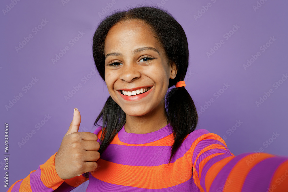 Close up happy little kid teen girl of African American ethnicity 15 years old wear striped orange sweatshirt do selfie shot pov mobile cell phone show thumb up isolated on plain purple background..