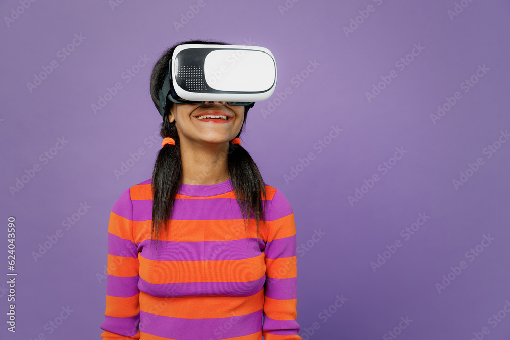 Little fun kid teen girl of African American ethnicity 15-16 years old wear striped orange sweatshirt watching in vr headset pc gadget isolated on plain purple background. Childhood lifestyle concept. Little fun kid teen girl of African American ethnicity 15-16 years old wear striped orange sweatshirt watching in vr headset pc gadget isolated on plain purple background. Childhood lifestyle concept.