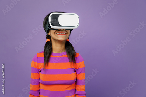 Little fun kid teen girl of African American ethnicity 15-16 years old wear striped orange sweatshirt watching in vr headset pc gadget isolated on plain purple background. Childhood lifestyle concept.