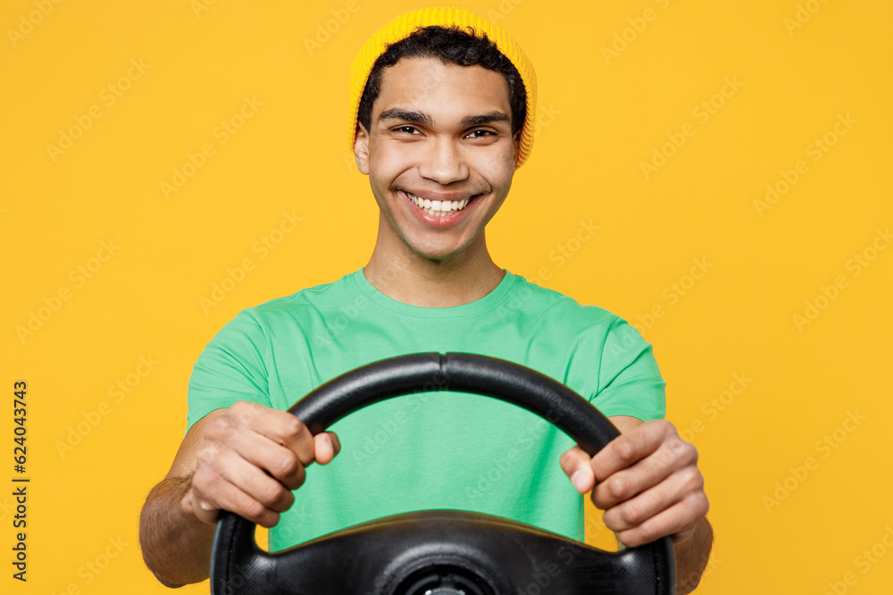 Young smiling happy man of African American ethnicity he wears casual clothes green t-shirt hat hold steering wheel driving car isolated on plain yellow background studio portrait. Lifestyle concept. Young smiling happy man of African American ethnicity he wears casual clothes green t-shirt hat hold steering wheel driving car isolated on plain yellow background studio portrait. Lifestyle concept.
