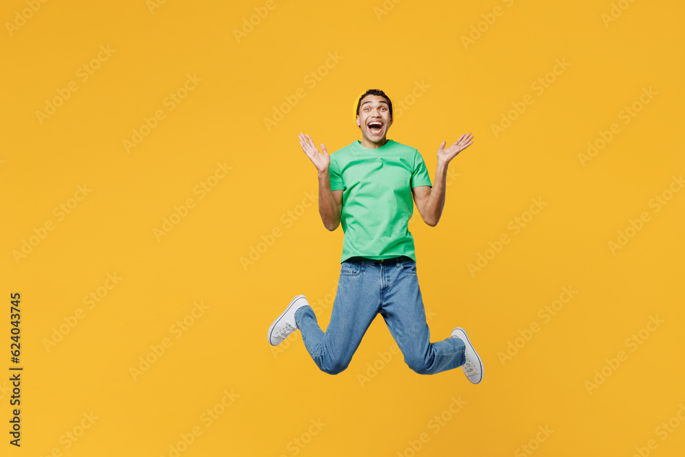 Full body young man of African American ethnicity he wears casual clothes green t-shirt hat jump high look camera spread hands isolated on plain yellow background studio portrait. Lifestyle concept.