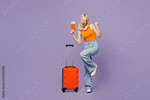 Traveler winner woman wears orange casual clothes hold suitcase passport ticket isolated on plain purple background. Tourist travel abroad in free time rest getaway. Air flight trip journey concept.