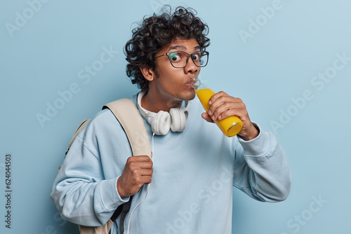 Horizontal indoor close up of young Hindu male wearing casual hoodie with white headphones on neck standing isolated in centre on blue background drinking delicious orange juice in glass bootle
