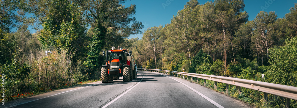 Tractor With Fertilizer Applicator With Tank In Motion On Country Road In Europe. Asphalt Road Against Background Of European Forest Landscape. Agricultural Concept