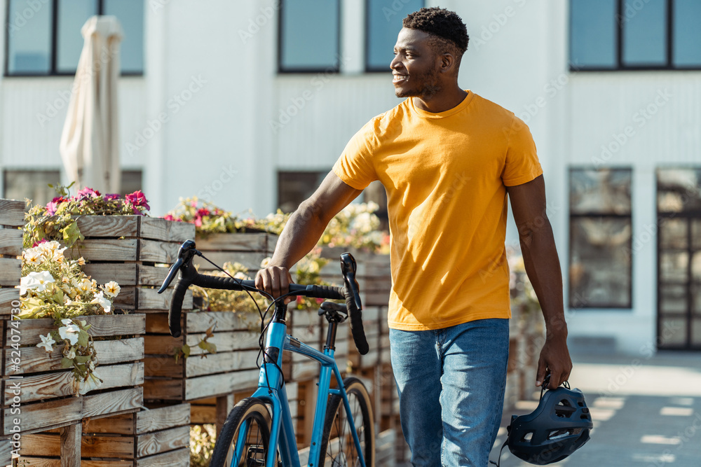 Positive smiling african man holding safety helmet walking with bicycle on street. Cycling concept