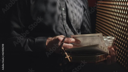 Medium section shot of unrecognizable Catholic priest holding rosary reading Bible or prayer book in confessional