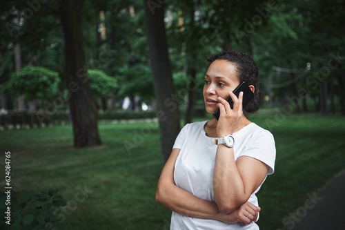 Confident multi-ethnic young pretty woman talks on mobile phone while strolling the alley of a city park on a summer day