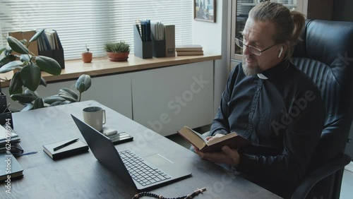 High angle shot of mature Catholic pastor sitting at desk in office checking something in book during online meeting with colleagues photo