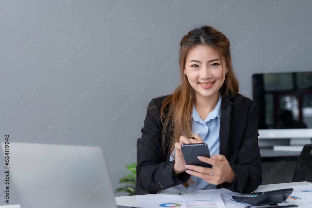 Portrait of a smiling asian businesswoman using mobile phone in the office. Young Asian businesswoman with paperwork at workplace using smartphone in a modern office. Portrait of a smiling asian businesswoman using mobile phone in the office. Young Asian businesswoman with paperwork at workplace using smartphone in a modern office.