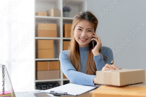 Young Asian business owner talking on smartphone and taking order. Asian female entrepreneur working at home office confirming the order on phone. © amnaj