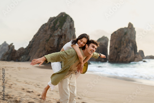 Beach adventure. Young man piggybacking his girlfriend and enjoying time by the ocean, free space #624124327