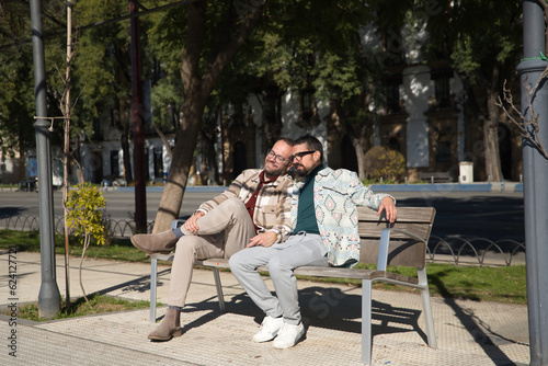 Real marriage of gay couple, holding hands, heads together and eyes closed, sitting on a wooden bench, complicit and happy. Concept lgtb, lgtbiq+, couples, in love, complicity.