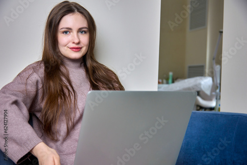 Young pretty student sits relaxed on a blue couch, holds a laptop, undergoes online training. Smiling freelance student using laptop to work remotely