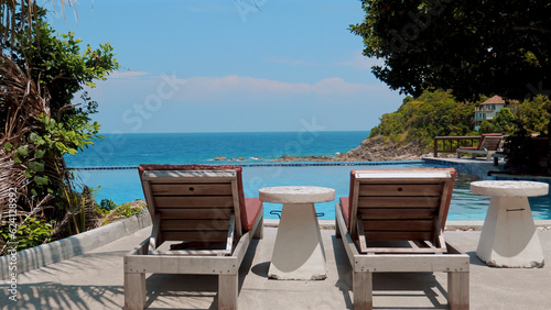 Wooden beach chairs and a table near a clear blue pool with sea view. Sunny day at a tropical hotel. Concept of summer vacation and relaxation.