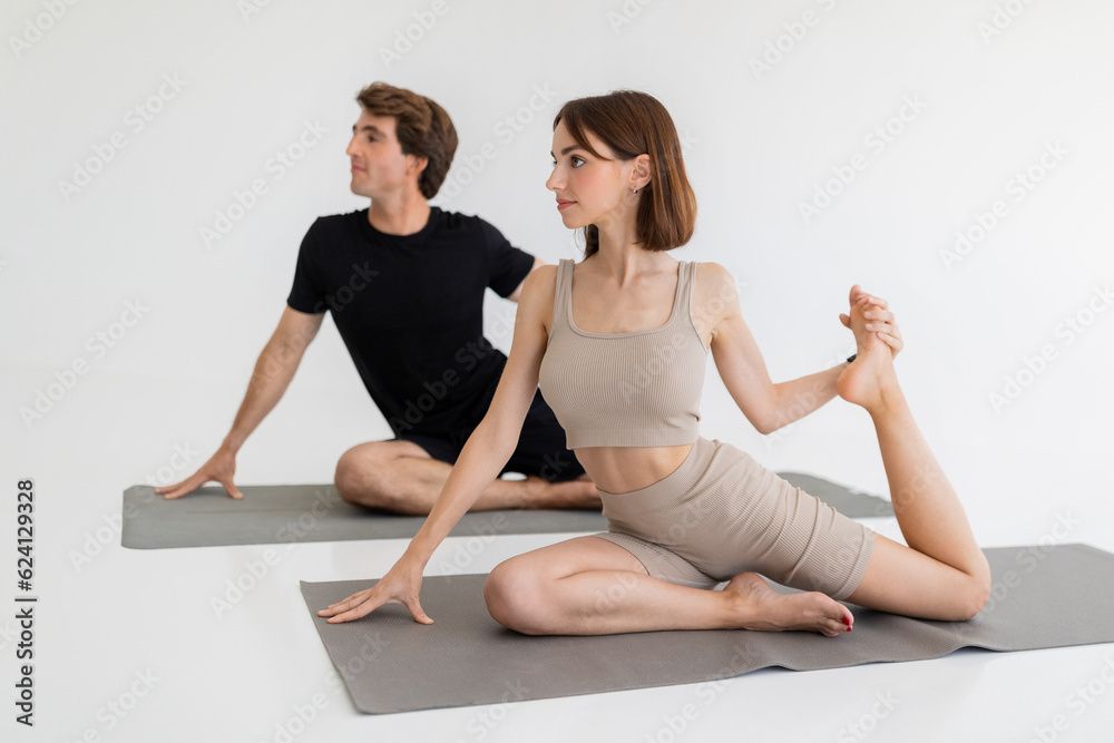 Calm millennial european couple in sportswear doing stretching exercise for legs, practicing yoga in gym Calm millennial european couple in sportswear doing stretching exercise for legs, practicing yoga in gym