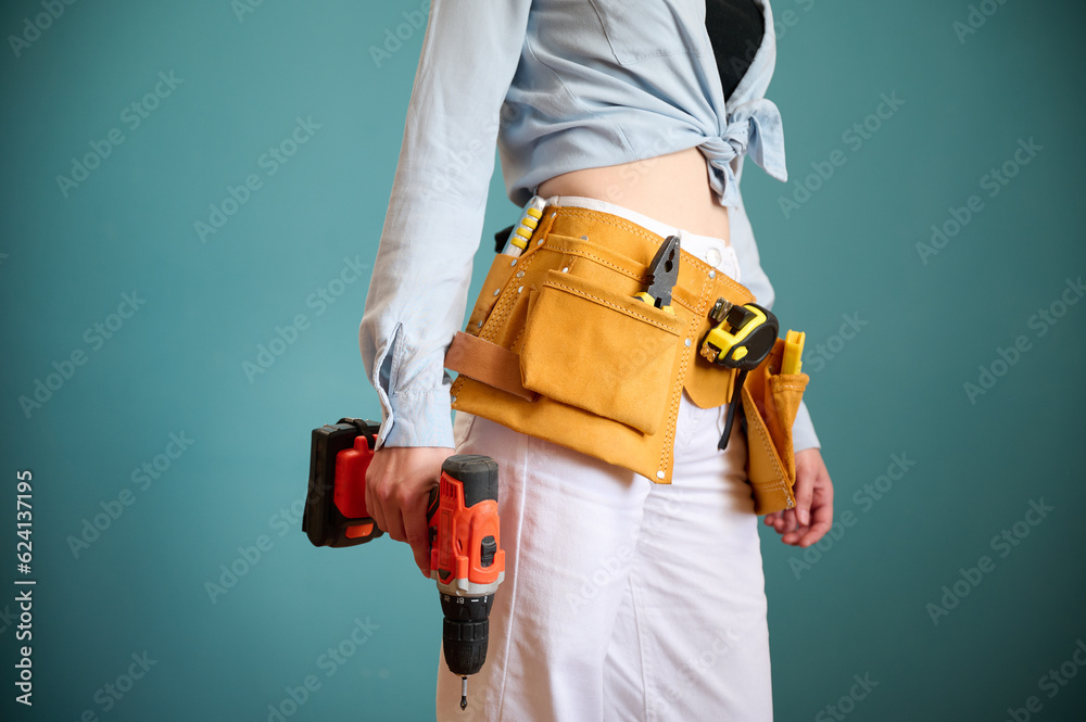 Young female worker with tool belt holding an electric screwdriver in hands isolated on background. Pretty caucasian female with tools planning new project. Portrait in studio. Crop view. Young female worker with tool belt holding an electric screwdriver in hands isolated on background. Pretty caucasian female with tools planning new project. Portrait in studio. Crop view.