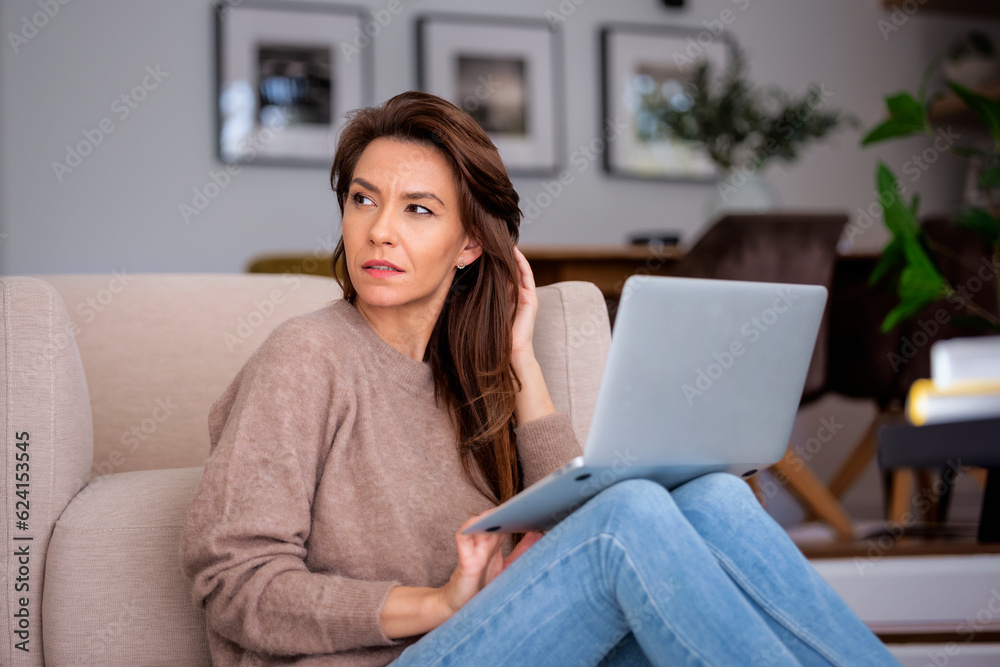 Naklejka premium Thinking caucasian woman sitting at home and using laptop