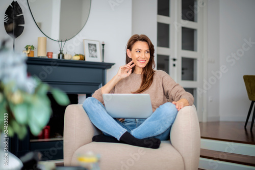 Thinking caucasian woman sitting in an armchair at home and using laptop