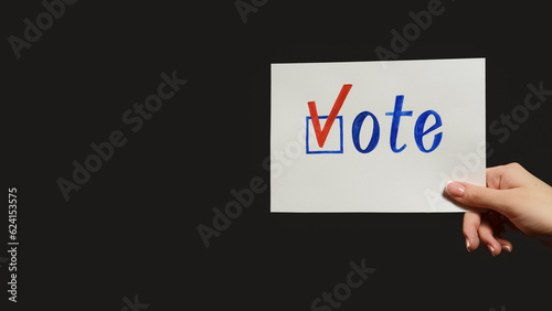 Vote sign. Election campaign. Political agitation. Female hand showing ballot with check mark isolated on dark black empty space background.
