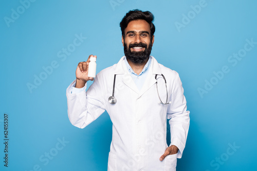 Positive middle aged indian man doctor in uniform holding jar of medicines, standing on blue background