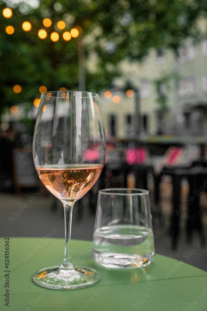 Obraz premium Glass of rose wine and water on a table of a city cafe terrace on summer evening. Street in bokeh.