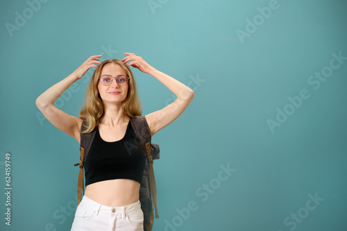 Young pretty female hipster with vintage backpack isolated on background. Attractive female student in glasses, short black top and white pants. Smiling woman showing expressions. Travel with backpack © Василь Івасюк