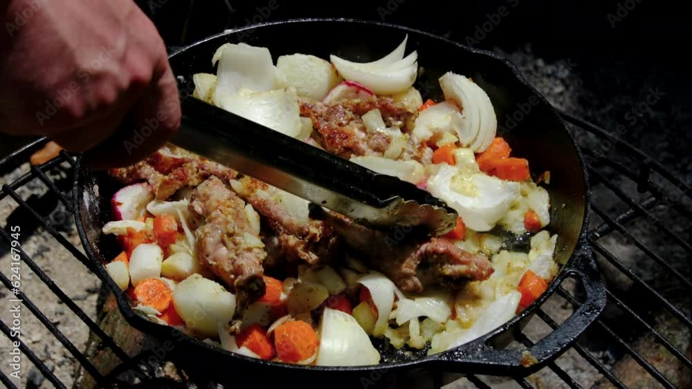 Using tongs to stir wild game meat being cooked over camp fire in cast iron skillet full of onions and carrots. Using tongs to stir wild game meat being cooked over camp fire in cast iron skillet full of onions and carrots.