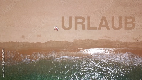 Luftaufnahme eines wunderschönen Sandstrandes am blauen Meer, an dem zwei Urlauber liegen und sich sonnen, daneben der Schriftzug „URLAUB“ im Sand