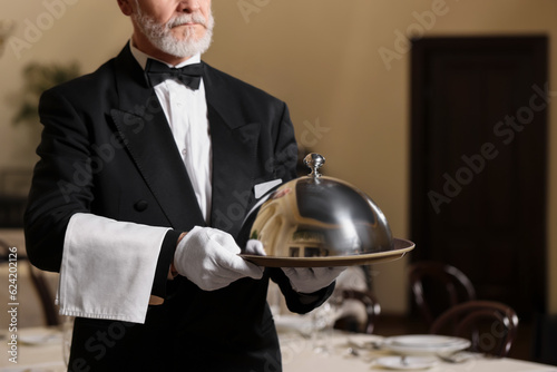 Butler holding metal tray with lid in restaurant, closeup. Space for text photo