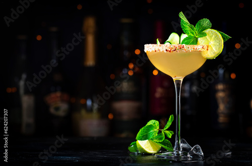 Cucumber margarita alcoholic cocktail drink with tequila, lime juice, syrup, mint and ice in glass garnished with salted chili pepper rim, dark background, bar tools and bottles
