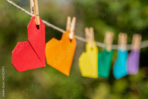 close em origami de coração vermelho pendurado por um mini pregador de madeira em varal, seguido por vários outros origamis de corações coloridos e desfocados em homenagem ao movimento LGBTQIAPN+ com  photo