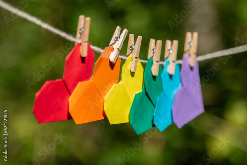 Varal de barbante com mini pregadores de madeira prendendo vários corações de origami coloridos nas cores da bandeira do orgulho gay em meio à natureza em fundo desfocado. photo