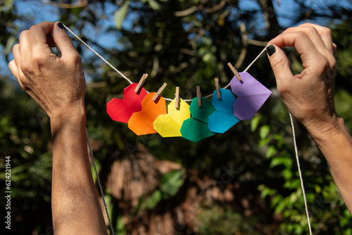 Mãos de mulher morena em meio à natureza pendurando um pequeno varal de corações de origami coloridos  simbolizando o movimento LGBTQIAPN+ photo