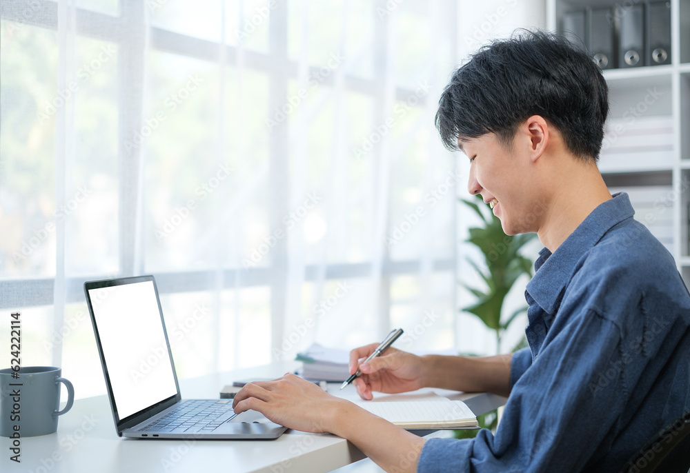 Satisfied young business man writing his strategy on paper and using laptop at office desk. Satisfied young business man writing his strategy on paper and using laptop at office desk.