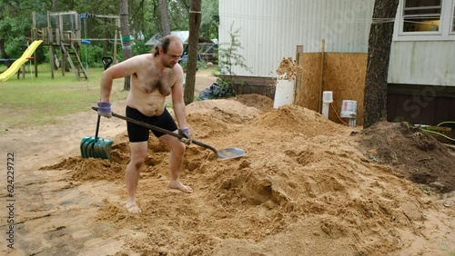 Shirtless and barefoot obese or man shoveling pile of sand into water well while wearing just briefs