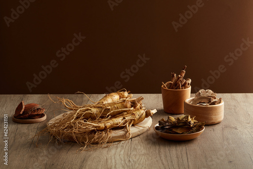 On the wooden table and brown background, chinese traditional medicine with fresh ginseng roots and some medicine herbs places on wooden plate and tray. Front view, herbs originating from the east