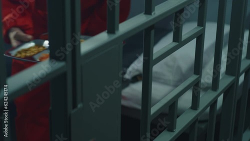 Close up of prison warden giving tray of food through metal bars to inmate. Elderly prisoner in orange uniform sits in prison cell, eats dinner. Serving imprisonment term in jail or detention center.