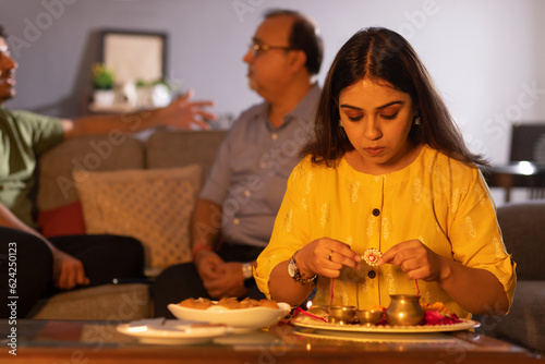 Young woman arranging puja thali on the occasion of Raksha Bandhan while her father and brother sitting behind
