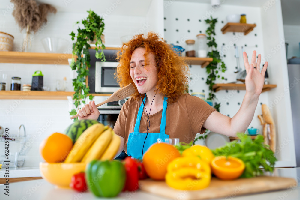 Funny beautiful woman singing into spatula, cooking in modern kitchen, holding spatula as microphone, dancing, listening to music, playful girl having fun with kitchenware, preparing food.